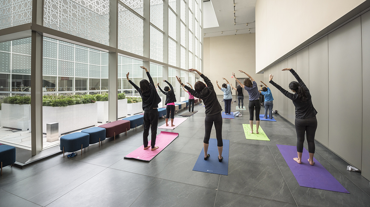 A group of women, facing away, lean to the left with arms raised during a yoga class in the Museum's atrium.